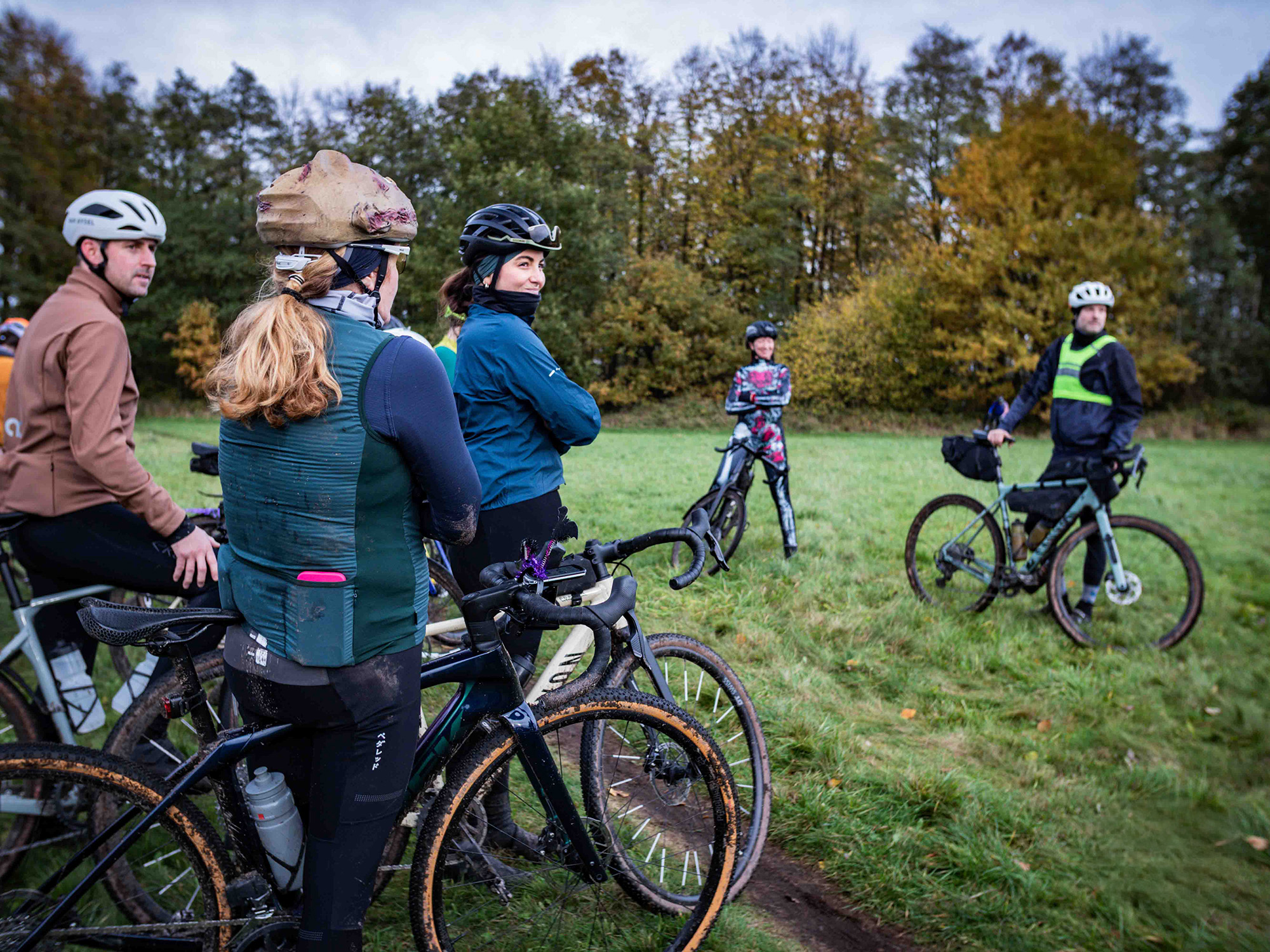 A group of five cyclists on gravel bikes standing in a grassy field. The two people in the foreground are looking off to the right, away from the camera. The woman in the center wears a teal jacket and has her arms crossed. The woman to her left is wearing a green vest over a dark long-sleeve top and has a distinctive helmet cover that looks like a hat with a brim. Their bikes are muddy. In the background, three other cyclists stand with their bikes; the one on the far right is wearing a dark jacket with a reflective green vest. Trees with autumnal foliage are visible behind them.
